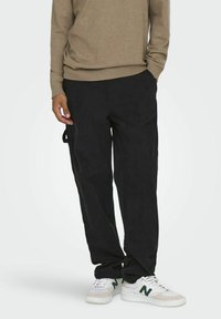 Sorte corduroy bukser med en løs pasform, sidelommer og en struktureret overflade, iført med en tan langærmet trøje og hvide sneakers.