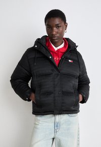 Black padded jacket with a hood, featuring a subtle logo pattern, button closure, and front pockets. Worn over a red hoodie.