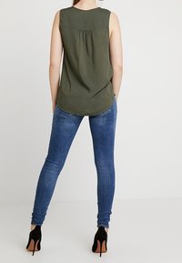 Olive green sleeveless blouse with a rounded hem and gathered back, paired with skinny blue jeans and black stiletto heels.