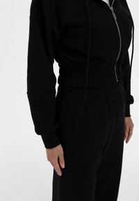 Mustat fleece-haalarit, joissa on vetoketjukiinnitys, röyhelöbändit ja kerääntynyt vyötärö, joka antaa istuvan ilmeen. Sujuva pinta koko vaatteen alueella.