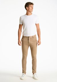 Pantalons slim beige avec une taille à bouton, associés à un t-shirt ajusté blanc. Le modèle se tient debout en baskets blanches sur un fond uni.