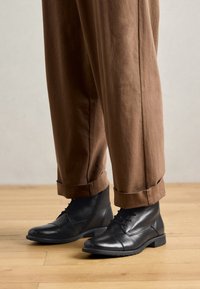 Schwarze Leder-Ankle-Boots mit runder Spitze, Schnürung vorne und strukturiertem Obermaterial. Kombiniert mit braunen, hochgekrempelten Hosen auf einem Holzfußboden.
