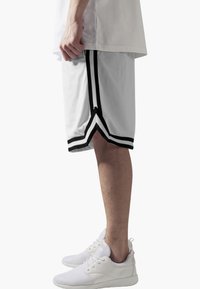 Shorts de basket en mesh blanc avec des rayures noires sur les côtés, présentant un détail d'ourlet en V. Associés à des baskets de sport blanches.