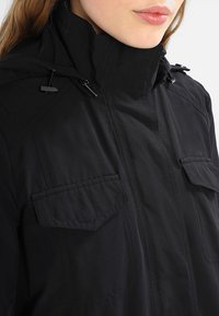 Chaqueta negra con capucha, cierre frontal, dos bolsillos con solapa, cordones ajustables en el cuello y tejido de textura suave. Diseño casual.