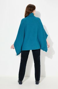 Poncho teal in maglia con colletto alto e maniche ampie. Indossato sopra pantaloni in velluto a coste neri, mostrando una vestibilità ampia e rilassata.