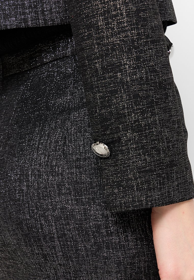 Blazer noir texturé avec détails en fil métallique, présentant un accent de bouton argenté sur la manche. Coupe ajustée et design structuré.