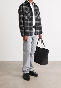 Schwarz-grün kariertes Hemd mit zwei Brusttaschen, graue Jeans, schwarze Lederschuhe und eine schwarze Tragetasche. Lässiges Outfit vor neutralem Hintergrund.