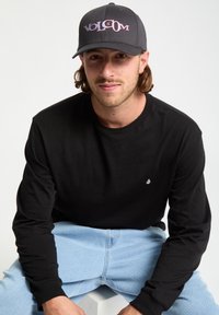 Harmaa lippis, jossa on punaisella ja valkoisella brodeerattu "Volcom", musta pitkähiha, vaaleansiniset farkut, istuu yksinkertaista taustaa vasten.