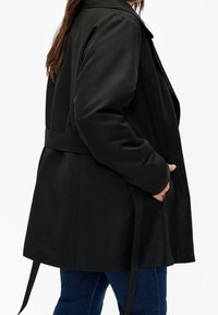 Svart ytterrock med skärp i midjan, med krage och sidofickor. Slät textur och något oversize passform.