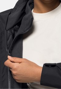 Musta vetoketjullinen takki pehmeällä fleecevuorilla, jossa on korkea kaulus ja säädettävät hihansuut. Kankaan pinta on sileä.
