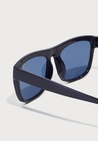 Ochelari de soare cu rame din plastic de culoare bleumarin, lentile nuanțate în albastru, formă rectangulară și brațe late. Prezintă o textură netedă și nu au hardware vizibil.