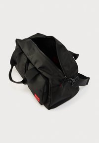 Μαύρη υφασμάτινη τσάντα με μεγάλο άνοιγμα φερμουάρ, διπλές κοντές λαβές, ρυθμιζόμενο ιμάντα και μικρή κόκκινη ετικέτα λογότυπου στο μπροστινό μέρος.