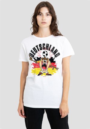 Junge Frau trägt ein weißes T-Shirt mit der Zeichentrickfigur Tasmanischer Teufel, einem Fußball und dem Wort Deutschland über den Farben der deutschen Flagge.