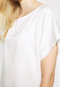 Biała, lekka bluzka z krótkimi, podwiniętymi rękawami. Materiał ma miękką teksturę i luźny krój, z prostym okrągłym dekoltem.