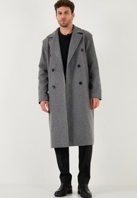 Uomo che indossa un lungo cappotto grigio doppiopetto, camicia nera, pantaloni neri e stivali in pelle nera su uno sfondo bianco semplice.