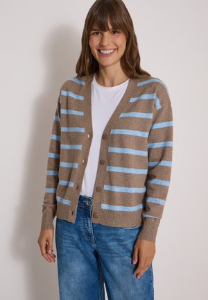 Femme souriante portant un cardigan marron avec des rayures horizontales bleu clair, une chemise blanche et un jean bleu sur un fond clair uni.