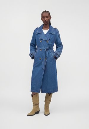 Kvinde iført en lang blå denim trenchcoat med bælte, kombineret med beige støvler til midt på læggen, stående mod en ensfarvet baggrund.