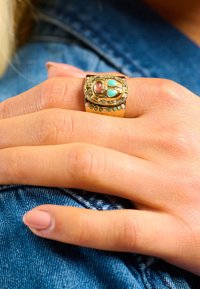 Bague dorée présentant un design de scarabée avec des accents turquoise et rose, montée sur un anneau lisse agrémenté de petites pierres colorées.