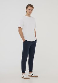 T-shirt bianco a maniche corte con scollatura rotonda, abbinato a pantaloni della tuta tapered blu navy. Il modello indossa sneakers bianche con accenti marroni.
