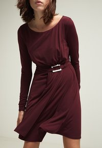 Bordowa dopasowana sukienka z długimi rękawami, okrągłym dekoltem i skręconym przodem. Posiada akcent w postaci klamry w srebrnym odcieniu na talii.