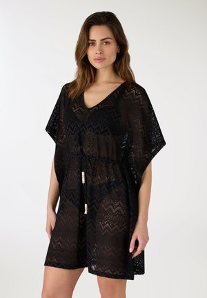 Donna che indossa un abito in pizzo trasparente nero con motivo a zigzag, scollo a V e vita con coulisse, in piedi davanti a uno sfondo chiaro e uniforme.