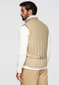Hombre de perfil que lleva un chaleco acolchado beige sobre un suéter blanco de cuello alto y tejido de ochos y pantalones beige frente a un fondo liso.