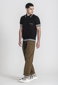 Černé polo tričko s texturovaným vzorem, bílé pruhy na rukávech a kapesníček na hrudi. V kombinaci s hnědými kalhotami s volnějším střihem a vzorovanými teniskami.