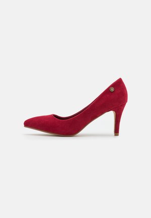 Escarpin en daim rouge à talon moyen, avec bout pointu et petit détail métallique rond près du talon, présenté sur fond blanc.
