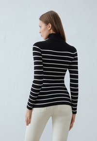 Svart og hvit stripete turtleneck-genser med en tettsittende design og ribbestruktur. Iført lyse jeans, sett fra bak.