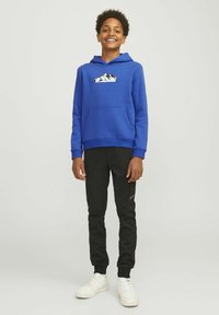 Blauer Hoodie mit einer Fronttasche und einem Berg-Graphic, kombiniert mit schwarzen tapered Jogginghosen. Weiße Sneakers runden das Outfit ab.