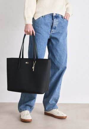 Bolsa tote de couro preto texturizado com alças longas verdes, apresentando um detalhe em dourado de chaveiro, segurada por uma pessoa vestindo denim azul claro.