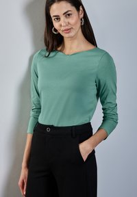 Vihreä pitkähihaista ykköstä V-pääntiellä, pehmeästä kankaasta valmistettu, yhdistetty korkeavyötäröisiin mustiin housuihin, joissa on etutasku ja nappidetalji.