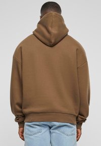 Brauner Kapuzenpullover aus dickem Stoff mit lockerer Passform. Verfügt über eine große Kapuze auf dem Rücken und gerippte Bündchen, kombiniert mit hellblauen Jeans.