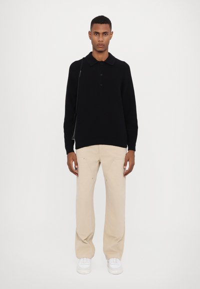 Polo shirt noir à manches longues avec trois boutons et un col, associé à un pantalon large beige et des sneakers blanches. Design simple et épuré.