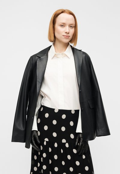 Μαύρο δερμάτινο μπουφάν, λευκό πουκάμισο με πέρλες στον γιακά, μαύρη φούστα με πουά, και μαύρα δερμάτινα γάντια. Ομαλή υφή, εφαρμοστό κόψιμο.