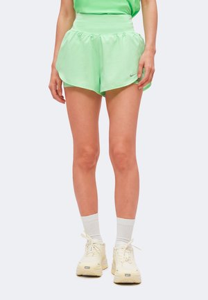 Mujer con pantalones cortos deportivos verde claro, calcetines blancos y zapatillas Nike color crema, de pie frente a un fondo liso.
