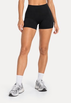Mujer con pantalones cortos deportivos negros de talle alto, calcetines blancos, y zapatillas de correr grises New Balance, de pie sobre fondo blanco.
