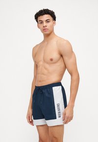 Morske kopalne hlače iz lahkega materiala, z belimi stranskimi paneli in logotipom "CALVIN KLEIN". Elastičen pas z vrvico.