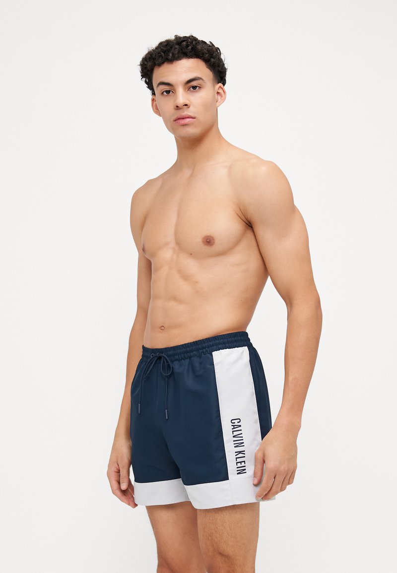 Morske kopalne hlače iz lahkega materiala, z belimi stranskimi paneli in logotipom "CALVIN KLEIN". Elastičen pas z vrvico.