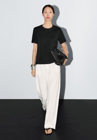 T-shirt nero a maniche corte, pantaloni bianchi a gamba larga e sandali neri con cinturini. La modella porta una borsa a tracolla nera con texture e indossa un bracciale d'argento.