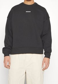 Personne portant un sweat-shirt noir à col rond avec le petit texte blanc « WRSTBHVR » centré sur la poitrine et un pantalon beige clair.