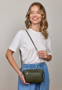 Zelená kožená taška přes tělo s hladkým povrchem, obsahuje přední kapsu na zip a nastavitelný popruh. Kompaktní a obdélníkový tvar.