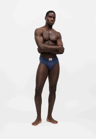 Homme musclé debout pieds nus, bras croisés, portant un slip bleu sur fond blanc uni.