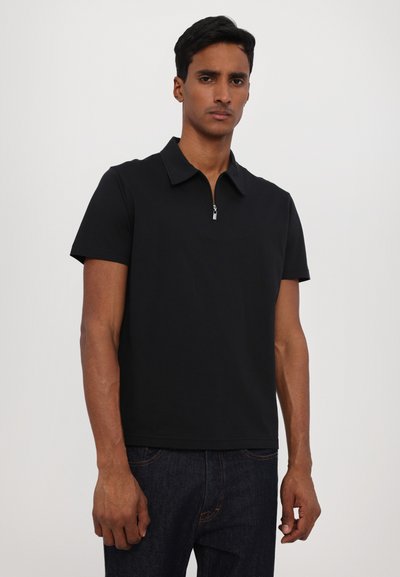 Polo preto de mangas curtas com gola com fecho éclair, corte reto, feito de um tecido liso e leve. Usado com jeans escuros.