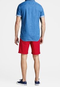 Se ve a una persona de espaldas, vistiendo una camisa de mezclilla de manga corta, pantalones cortos rojos y zapatillas azul marino, de pie contra un fondo liso.