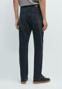 Persona che indossa jeans dritti blu scuro e scarpe marroni, in piedi contro uno sfondo chiaro e uniforme, vista da dietro.