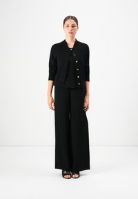Kvinna som står i en svart knappad cardigan med hjärtformade knappar, vida svarta byxor och svarta öppna skor på vit bakgrund.