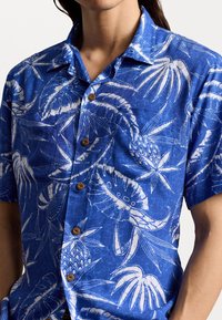 Koszula z krótkim rękawem zapinana na guziki w niebieskiej tkaninie z białymi wzorami tropikalnymi, z klasycznym kołnierzykiem i brązowymi guzikami.