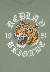 Zelené tričko s velkým grafickým motivem tygří hlavy s červenýma očima, obklopené textem "REPLAY BRIGADE 1981" v bílé barvě.