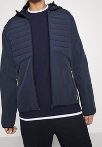 Marinblå jacka med huva och dragkedjestängning. Har vadderade paneler på axlarna, två dragkedjeförsedda framfickor och textil med struktur.